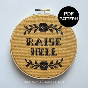 May include: A yellow cross stitch hoop with black embroidery that reads "Raise Hell" with floral accents at the top and bottom.