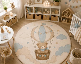 Op de afbeelding: Een rond vloerkleed met een centrale illustratie van een bruine teddybeer in een heteluchtballon. Het ontwerp bevat lichtblauwe en beige wolken, sterren en vlaggetjes op een crèmekleurige achtergrond met een afgewerkte rand.