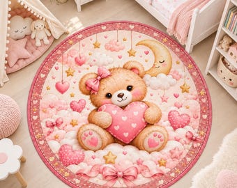 Op de afbeelding: Een rond vloerkleed met een lichtbruine teddybeer in het midden, voorzien van een roze strik op het hoofd, die een groot roze hart met witte hartjes vasthoudt. De achtergrond toont roze wolken, hangende sterren, hartjes en een maan.