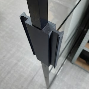 Op de afbeelding: Een matzwarte, 3D-geprinte kunststof beugel met een centraal rechthoekig kanaal en twee zijdelingse groeven. Het onderdeel is stevig bevestigd op een dunne, verticale zwarte metalen staaf en heeft een getextureerde afwerking en stijve constructie.