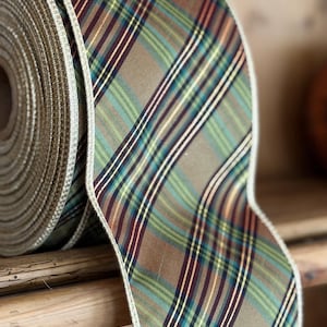May include: A roll of wide fabric ribbon featuring a plaid pattern in shades of olive green, brown, dark blue, and thin white stripes. The ribbon has a metallic gold finished edge and a textured weave, shown coiled on a wooden surface.