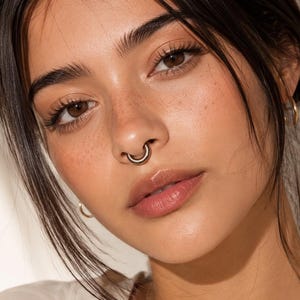 May include: A gold septum ring featuring a smooth, circular hoop design worn in the nose. The jewelry is crafted from a polished metallic material with a simple, continuous band that curves to fit the septum area, providing a clean look.