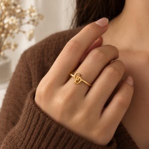 May include: A gold-toned ring featuring a stylised monogram design with the letters C and D intertwined. The ring has a thin, polished band and is worn on a finger. The metal has a smooth, reflective finish that catches the light clearly.