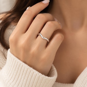 Könnte beinhalten: Ein Ring aus poliertem Silber mit einem glatten, durchgehenden wellenförmigen Design, das sich sanft entlang der Ringschiene krümmt. Das Metall hat eine reflektierende, hochglänzende Oberfläche, die ein schlichtes Erscheinungsbild erzeugt.