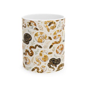 May include: A white ceramic mug featuring a repeating pattern of various ball pythons in shades of orange, yellow, brown, and black. The design includes small botanical sprigs and tiny gold stars scattered across the surface of the mug.
