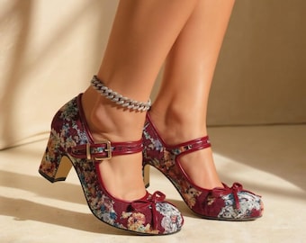 May include: A pair of Mary Jane heels featuring a dark red floral tapestry pattern, a rounded toe with a small bow, a buckled strap across the instep, and a block heel. The shoes are accented with solid dark red trim along the edges and straps.