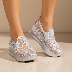 May include: A pair of silver slip-on sneakers featuring a sheer mesh upper with an intricate, swirling filigree pattern embellished with small, multicolored rhinestones. The shoes have a solid silver platform sole and a rounded toe design.