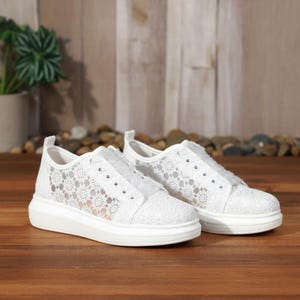 May include: A pair of white sneakers featuring a delicate floral lace overlay on the sides and toe box. These low-top shoes have a thick white rubber sole, white laces, and a structured heel tab, providing a clean and uniform appearance.
