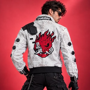 May include: A light gray distressed jacket featuring a large red and black graphic of a stylized demon mask on the back. The collar displays the text SAMURAI in white letters. The sleeves have black circular accents, and the hem is black.