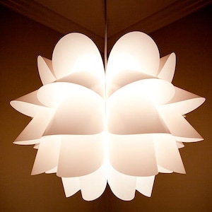 Könnte beinhalten: Eine weiße Pendelleuchte mit einem kugelförmigen, mehrschichtigen Design aus überlappenden, geschwungenen Blütenblättern. Die Lampe strahlt ein weiches, warmes Licht von innen aus und betont die durchscheinenden Kanten der an einem Kabel hängenden Struktur.