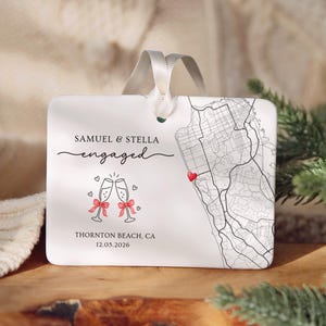 May include: A rectangular white ceramic ornament with a white ribbon hanger. It features the text Samuel & Stella engaged, a graphic of two champagne flutes with red bows, a black line map of Thornton Beach, CA, a red heart, and the date 12.05.2026.