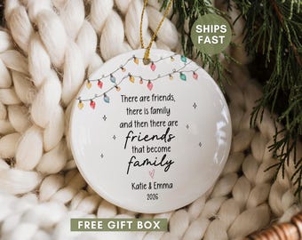 May include: A round white ceramic ornament featuring a string of colorful Christmas lights at the top and the text: There are friends, there is family and then there are friends that become family, Katie & Emma, 2026, with a small pink heart.