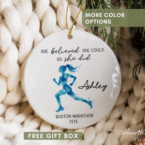 May include: A round white ceramic ornament featuring a blue watercolor graphic of a runner. The text reads: She believed she could. so she did, Ashley, and Boston Marathon 2026. The ornament hangs from a gold string.