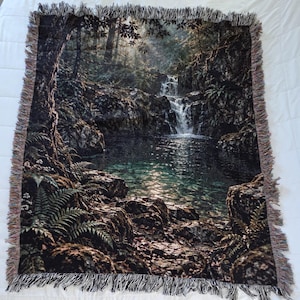 May include: A rectangular woven throw blanket featuring a detailed landscape scene of a waterfall cascading into a clear pool surrounded by dark rocks, lush green ferns, and dense forest trees, finished with a decorative fringed edge on all sides.