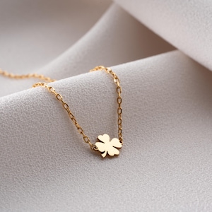May include: A gold-toned necklace featuring a delicate chain and a small, four-leaf clover charm. The metal has a smooth, polished finish, and the clover is centered along the chain, which is composed of small, uniform oval links.