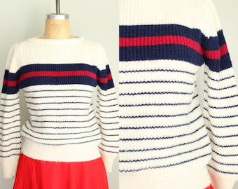May include: Cream-colored sweater with horizontal navy and red stripes across the chest. The sweater has long sleeves and a crew neck. The lower portion of the sweater features thin navy and cream stripes. The sweater is displayed on a mannequin.