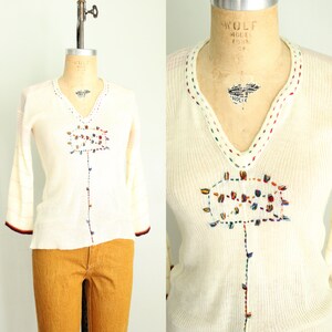 Peut inclure: Pull à manches longues de couleur crème avec un col en V et des détails brodés colorés. Le pull présente un motif floral sur le devant et des rayures sur les manches. Les poignets ont une bordure rouge foncé et marron.