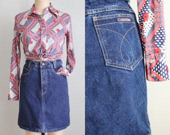 May include: A vintage denim skirt in a classic A-line silhouette. The skirt is a deep indigo blue with a single back pocket featuring the Calvin Klein logo. The skirt is paired with a patterned long-sleeve shirt in red, white, and blue.
