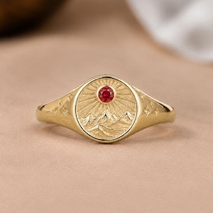 Puede incluir: Un anillo de sello de oro con una cara circular que presenta un paisaje montañoso grabado y un patrón de rayos de sol. Una pequeña piedra preciosa roja y redonda está engastada en el centro del sol. El aro tiene pequeñas estrellas grabadas.