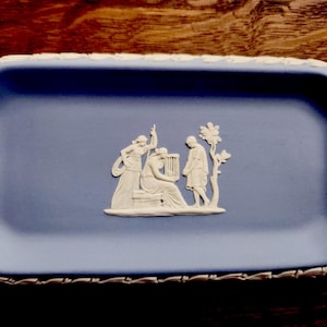 May include: A rectangular light blue ceramic tray featuring a white relief design in the center depicting three figures, one playing a harp, and a white decorative border pattern along the outer edge of the tray's rim.