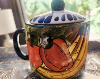 Può includere: Una tazza in ceramica con coperchio caratterizzata da un disegno dipinto a mano di mele rosse, banane gialle e un anacardo. La tazza presenta un manico scuro e un coperchio bianco decorato con motivi floreali di colore blu.