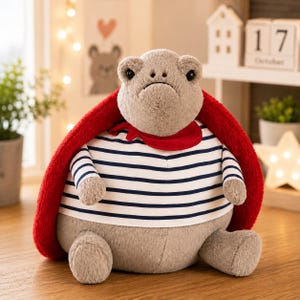 Puede incluir: Un peluche con forma de tortuga con un cuerpo suave de color marrón claro y un caparazón grande y rojo de textura afelpada. La tortuga lleva una camiseta blanca con rayas horizontales azul marino y un pañuelo rojo atado al cuello. Ojos oscuros.
