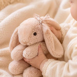 以下が含まれることがあります： 柔らかいライトベージュの毛並み、長い垂れ耳、片耳の付け根に結ばれた小さな繊細なサテンリボンの蝶結びが特徴のウサギのぬいぐるみ。このおもちゃは、暗い丸い目と縫い付けられた鼻を持ち、全体的にふわふわとした質感の外観をしています。