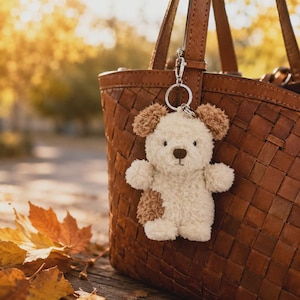 May include: A fluffy cream-colored plush bear keychain with brown ear accents and a brown patch on its side, attached to a silver metal ring. The bear is hanging from the handle of a brown woven leather handbag with a textured grid pattern.