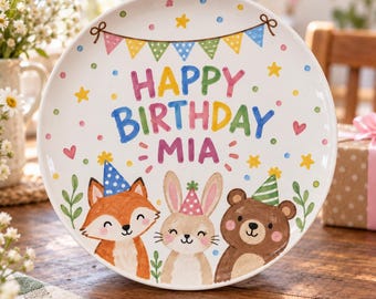 以下が含まれることがあります： パーティーハットをかぶったキツネ、ウサギ、クマのカラフルなイラストが描かれた白い陶器のプレート。中央には「HAPPY BIRTHDAY MIA」という文字が色とりどりのフォントでプリントされており、周囲には旗のガーランド、星、ドットが散りばめられています。