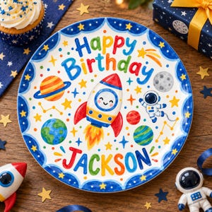 Peut inclure: Une assiette ronde en céramique avec un thème spatial coloré comprenant une fusée centrale, un astronaute, des planètes et des étoiles. Le texte Happy Birthday est imprimé en haut et le nom Jackson est inscrit en bas en lettres vives.