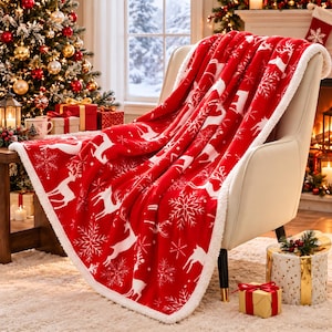 May include: A red throw blanket featuring a white pattern of reindeer and snowflakes, finished with a white sherpa fleece trim. The blanket is draped over a cream-coloured armchair, showcasing its soft texture and festive holiday design.