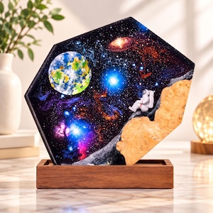 May include: A hexagonal resin art piece featuring a deep space nebula scene with a glowing planet, bright stars, and a small astronaut figurine perched on a natural wood edge, all mounted on a rectangular wooden base stand.
