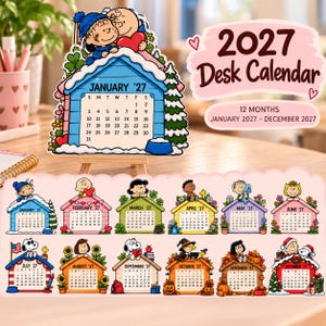 May include: A 2027 desk calendar featuring Peanuts characters on each monthly page. The calendar displays 12 months from January to December 2027, with each month illustrated by a different character scene set atop a house-shaped calendar grid.