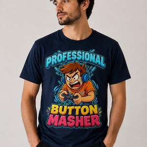 May include: A navy blue short-sleeve t-shirt featuring a graphic illustration of a cartoon character wearing blue headphones and playing a video game. The text "PROFESSIONAL BUTTON MASHER" is printed in bold, colorful letters above and below the character.