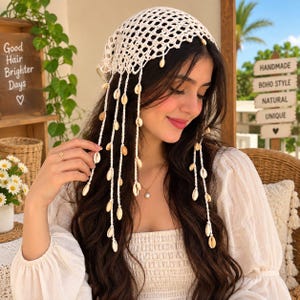 May include: A cream-colored crochet head scarf featuring an open-knit mesh pattern and long, dangling fringe strands adorned with small, natural white cowrie shells. The head covering is designed to tie at the back with matching crochet strings.