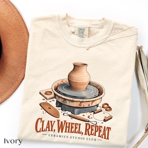 May include: An ivory t-shirt featuring a graphic of a pottery wheel with a clay vase, sculpting tools, and the text "CLAY, WHEEL, REPEAT CERAMICS STUDIO CLUB" in brown. The shirt has a Comfort Colors brand label at the inner neckline.