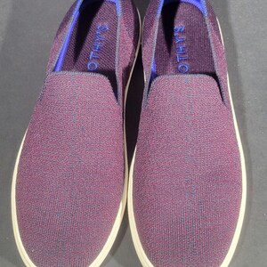 May include: A pair of slip-on shoes featuring a textured, deep purple knit fabric upper. The interior collar is lined with a bright blue trim, and the insole displays the text ROTHY'S in blue. The shoes are finished with a solid white rubber sole.