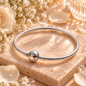 May include: A polished silver bangle bracelet featuring a spherical clasp engraved with the text ORA DOKA. The bracelet has a smooth, circular band design with two small ribbed accents flanking the central spherical closure piece.