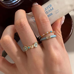 Op de afbeelding: Drie goudkleurige ringen gedragen aan de vingers. Eén ring heeft kleine bloemvormen met lichtblauw en wit emaille. Een andere is een dunne gouden band met kleine blauwe en doorzichtige steentjes. De derde is een gladde lichtblauwe emaille ring met steen.