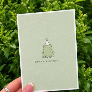 May include: A rectangular greeting card with a light sage green background featuring a central illustration of a small green frog wearing a polka-dot party hat. Below the frog, the text reads HOPPY BIRTHDAY. in a simple black sans-serif font.