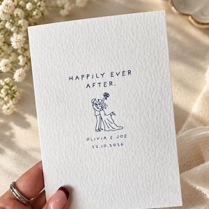 May include: A white textured card featuring dark blue printed text that reads HAPPILY EVER AFTER. Below the text is a line art illustration of two figures embracing, with the names OLIVIA & JOE and the date 22.10.2026 printed underneath.
