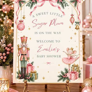 May include: A rectangular baby shower sign featuring a pink nutcracker and a mouse ballerina in a tutu. The text reads A SWEET LITTLE Sugar Plum IS ON THE WAY WELCOME TO Emilia's BABY SHOWER, decorated with pink ribbons, pine branches, and gifts.