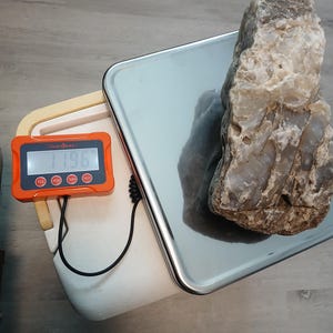 May include: A large, rough-textured, translucent gray and white rock sits on a silver digital scale. The orange display unit, connected by a black cord, shows a weight reading of 11.96 pounds. The scale features a flat, reflective metal platform.