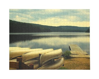Könnte beinhalten: Ein Puzzle, das eine Landschaft mit mehreren hellen Kanus zeigt, die auf einem Holzgestell im Vordergrund gestapelt sind. Ein Holzsteg ragt in einen ruhigen See hinein, wobei im Hintergrund eine dunkle, bewaldete Bergkette unter einem bewölkten Himmel zu sehen ist.