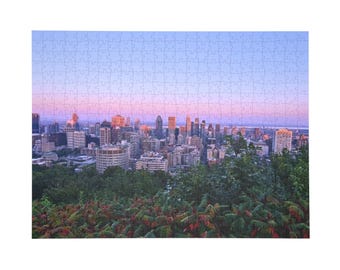 Könnte beinhalten: Ein Puzzle mit einer Panoramaansicht einer Stadtsilhouette bei Sonnenuntergang. Die Szene zeigt zahlreiche Gebäude und Wolkenkratzer unter einem Farbverlaufshimmel von Hellblau bis zartem Rosa, mit grünem Blattwerk im Vordergrund.
