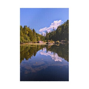 May include: A jigsaw puzzle featuring a landscape of a calm lake reflecting a clear blue sky with white clouds. Dense green forests line the shore, with large rocks visible both above the water and submerged near the foreground of the scene.