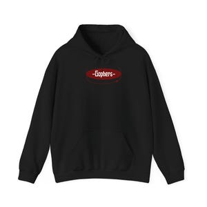May include: A black long-sleeve hooded sweatshirt featuring a front kangaroo pocket and a drawstring hood. Centered on the chest is a dark red oval graphic containing the white text -Gophers- with a thin red line circling the oval shape.