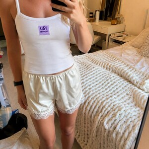 May include: A white tank top with thin straps featuring a purple rectangular graphic with the text UST TOMMIES on the chest, paired with cream-colored satin shorts that have a scalloped white lace trim along the bottom hem of each leg.