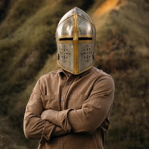 May include: A polished steel knight helmet featuring a gold-toned central cross bar and decorative gold-toned trim. The helmet displays engraved cross motifs on the front and a horizontal eye slit, worn with a plain tan long-sleeved button-down shirt.