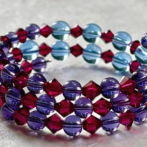 May include: A multi-strand wrap bracelet featuring alternating round light blue beads and faceted deep red bicone beads. The design includes a repeating pattern of translucent blue and dark red crystal-like elements arranged in a spiral coil structure.